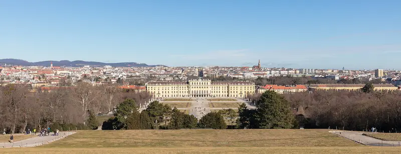 Palácio de Schönbrunn amarelo no fim do grande gramado dos jardins, visto da colina da Gloriette, com Viena e os morros ao fundo sob céu azul