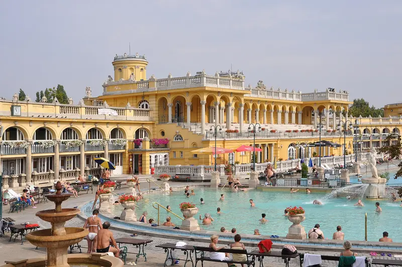 Piscina ao ar livre do Széchenyi com banhistas na água verde-azulada, diante dos prédios amarelos neobarrocos
