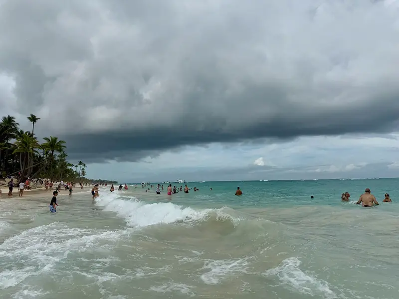 Ondas quebrando em Bávaro sob nuvens carregadas, com banhistas na água e coqueiros na faixa de areia à esquerda