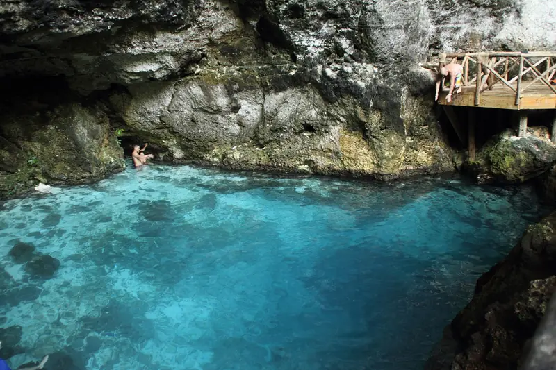 Poço de água azul-turquesa cercado de paredão de rocha, com deque de madeira à direita e banhistas na borda