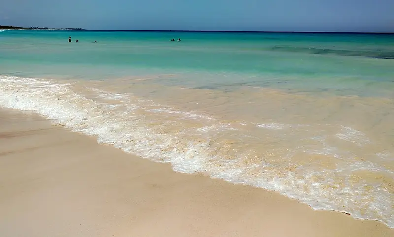 Faixa de areia clara com espuma de onda em primeiro plano, mar verde-claro e algumas pessoas na água ao fundo