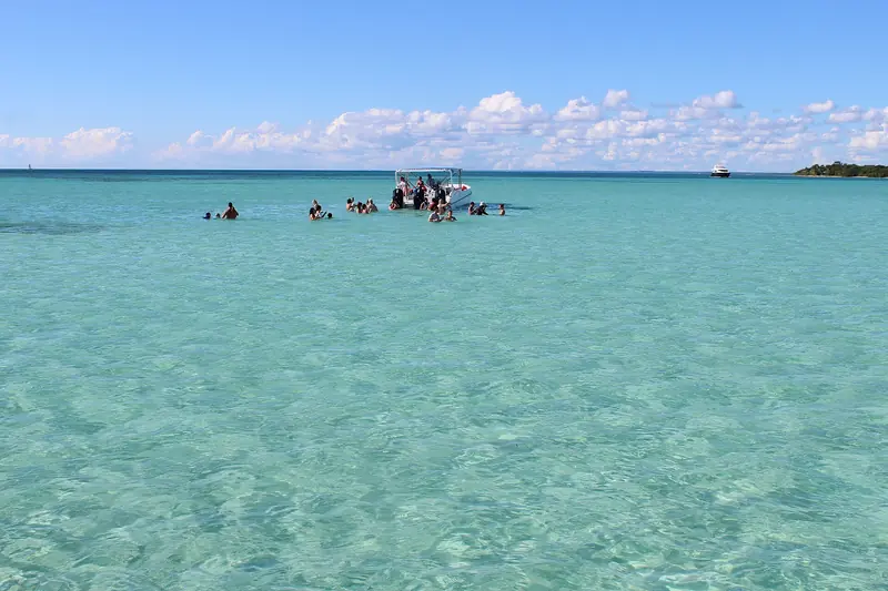 Grupo de turistas com água pela cintura ao redor de uma lancha num banco de areia de mar verde-claro, com ilha ao fundo