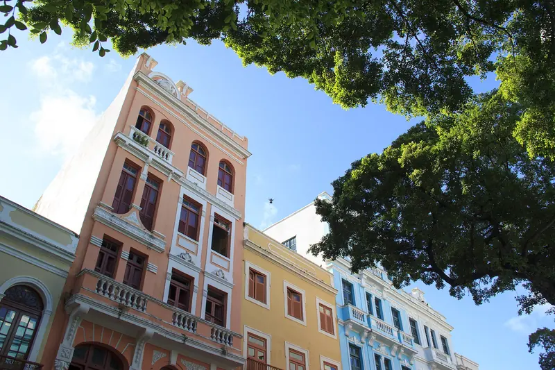 Fachadas de sobrados de três e quatro andares em tons de rosa, amarelo e azul, vistas de baixo, com a copa de uma árvore cobrindo parte do céu
