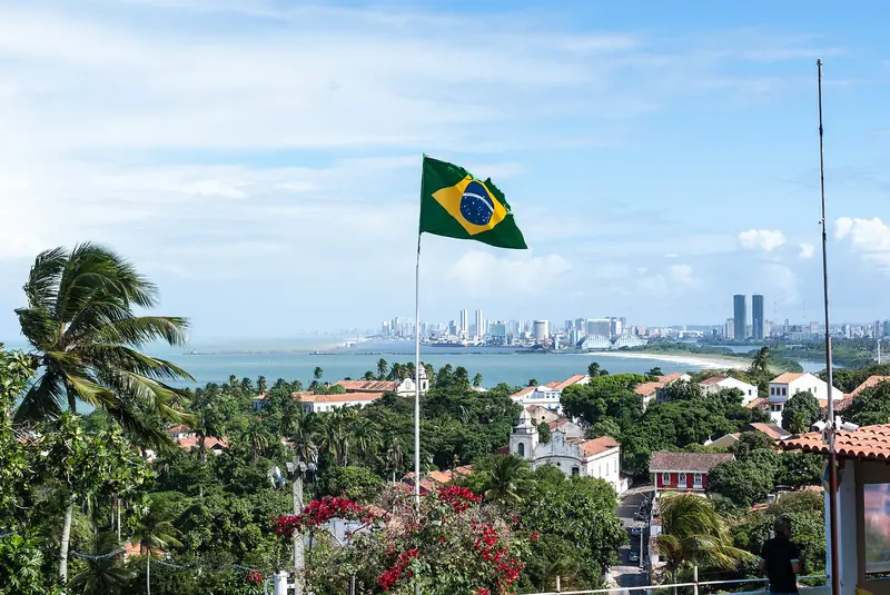 Vista do Alto da Sé de Olinda, com bandeira do Brasil, coqueiros, telhados e torres de igreja em primeiro plano e os prédios do Recife na linha do mar ao fundo