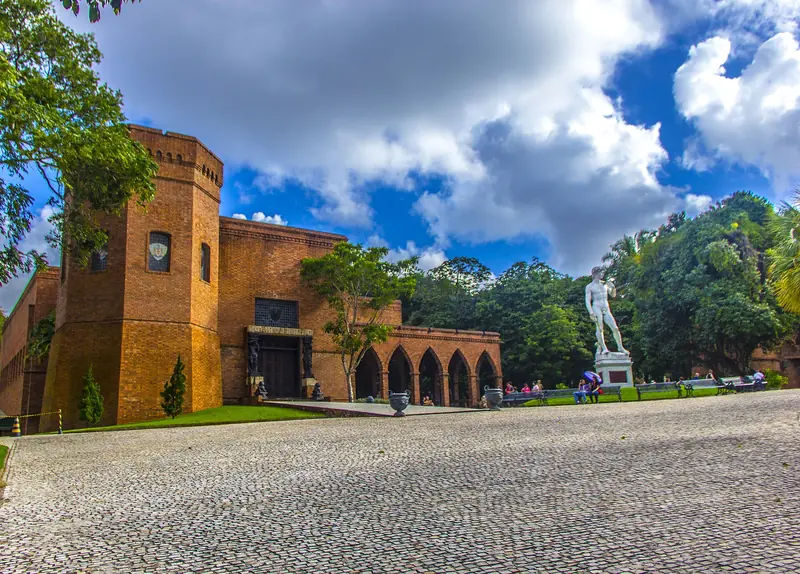 Castelo de tijolos alaranjados com torre e arcos góticos, diante de um pátio de pedras portuguesas, e uma estátua branca de mármore ao lado do jardim