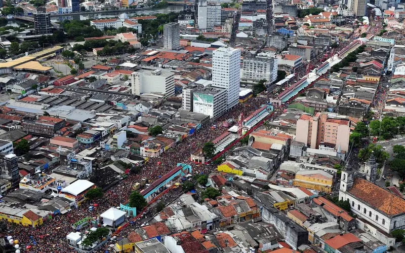 Vista aérea de uma avenida do centro do Recife tomada por uma multidão compacta entre prédios, com camarotes ao longo do percurso