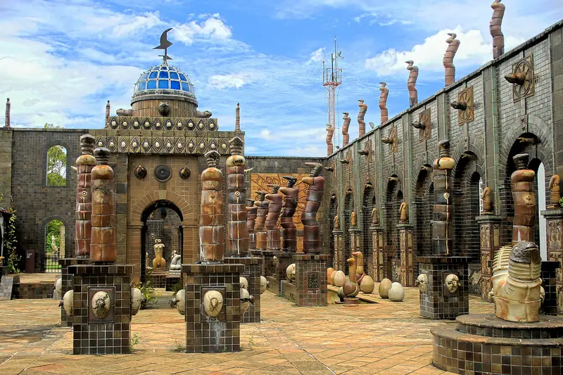Pátio de piso de cerâmica com totens de barro sobre pedestais, muros de tijolo escuro com arcos e uma cúpula de vidro azul ao fundo