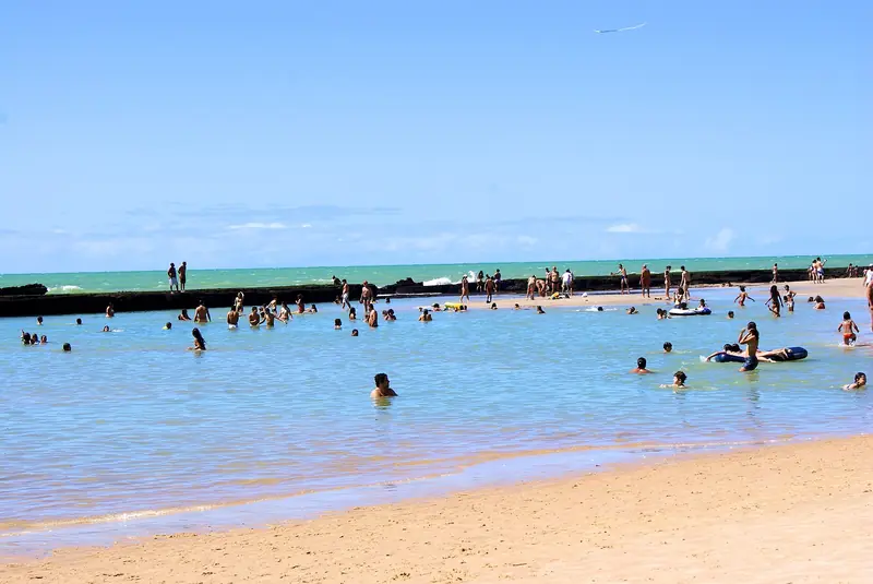 Banhistas numa piscina rasa e calma de água azul-clara, limitada ao fundo por uma faixa escura de recife, com o mar aberto verde depois dela