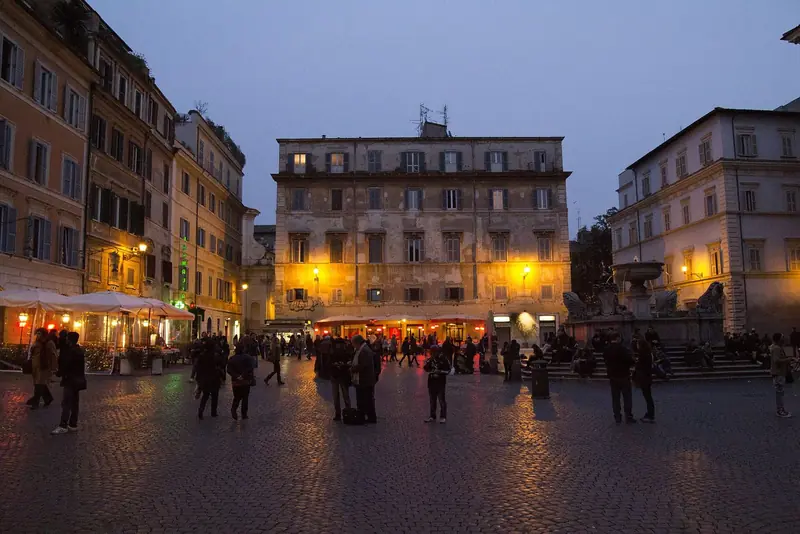 Piazza di Santa Maria in Trastevere ao anoitecer, com a fonte à direita e as luzes dos restaurantes acesas