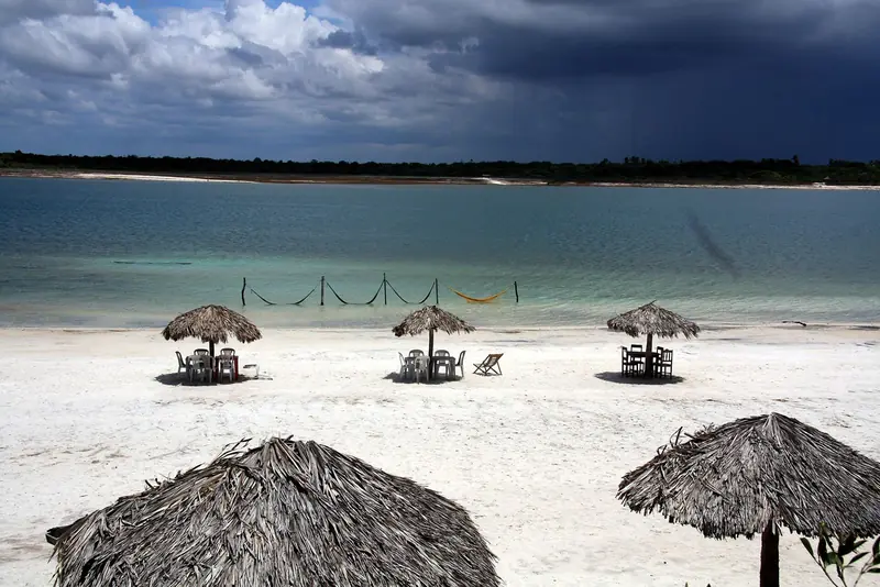 Areia branca com guarda-sóis de palha e redes armadas dentro da água azul-turquesa da Lagoa do Paraíso, sob céu carregado
