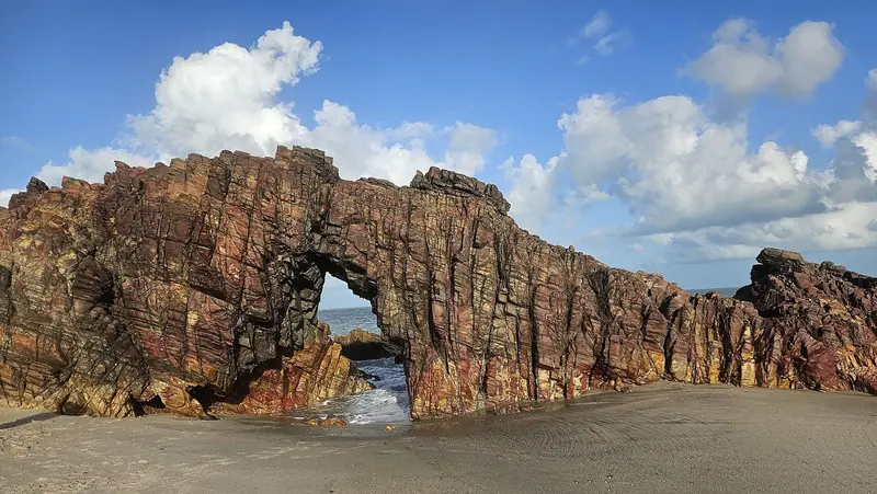 Arco de pedra avermelhada na areia da praia, com o mar visto através do furo e céu azul