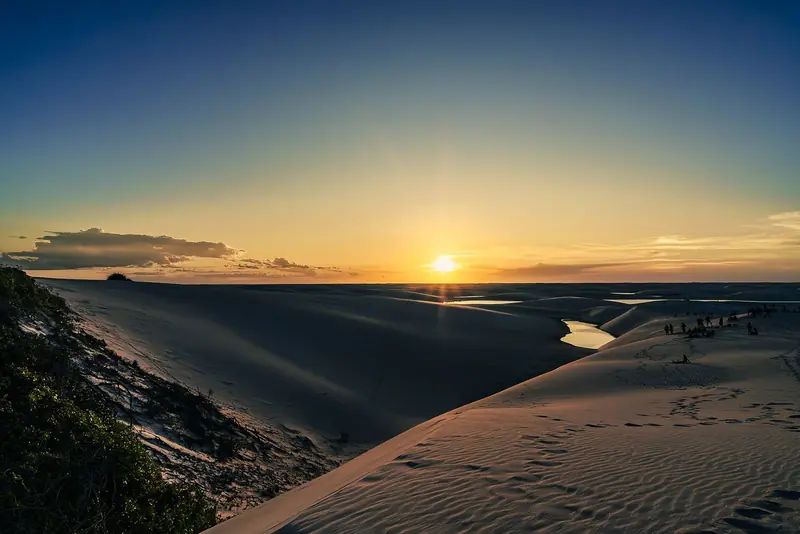 Pôr do sol visto do alto de uma duna, com lagoas entre as dunas e um grupo de pessoas ao longe