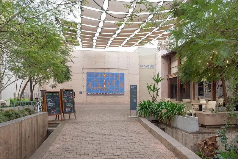 Pátio de pedra da praça Mulato Gil de Castro, com árvores, um toldo vazado e o mural de cerâmica azul na fachada do museu
