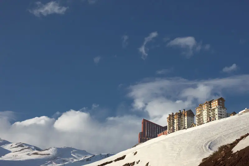 Prédios do Valle Nevado no alto de uma encosta nevada, sob céu azul com nuvens