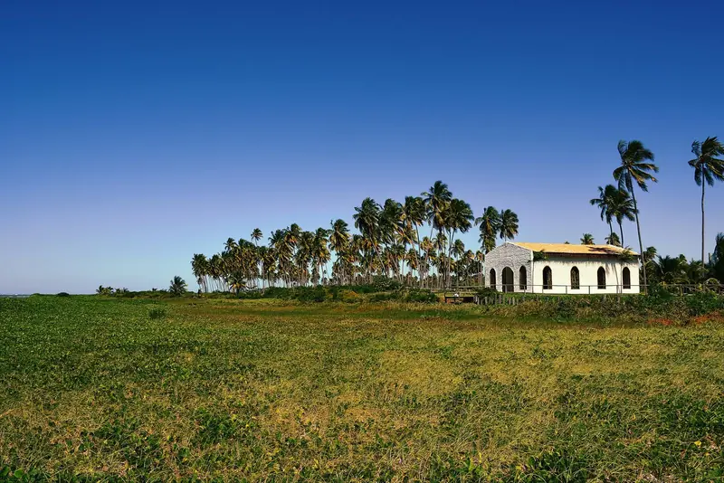 Pequena capela branca com arcos em meio a vegetação rasteira, com coqueiros ao fundo
