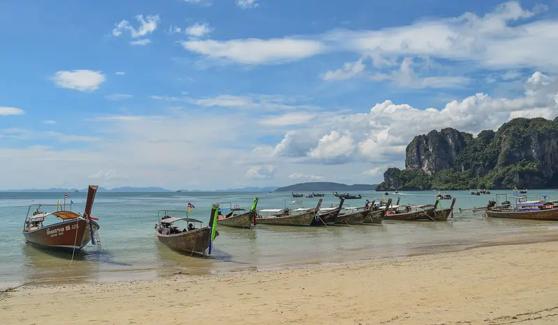 Barcos de cauda longa ancorados na areia clara de Railay Oeste, com o paredão de calcário coberto de mata à direita