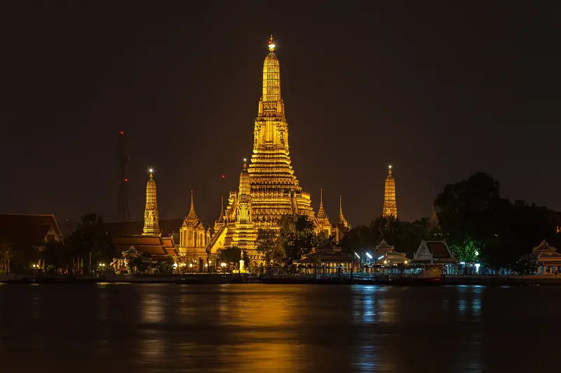 O Wat Arun iluminado à noite, com a torre central dourada e as torres menores refletidas no rio Chao Phraya