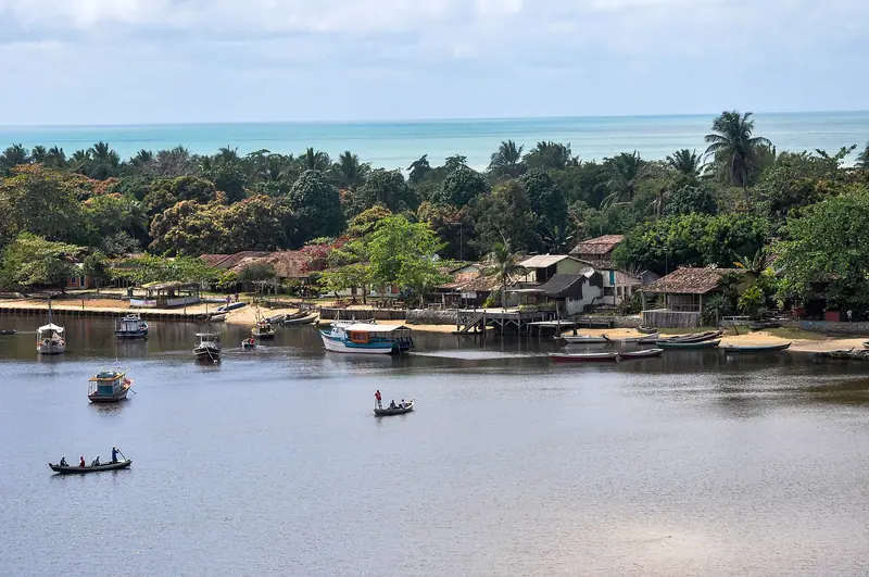 Vila de casas baixas e telhados de barro entre coqueiros, na margem de um rio largo com canoas e barcos, e o mar ao fundo