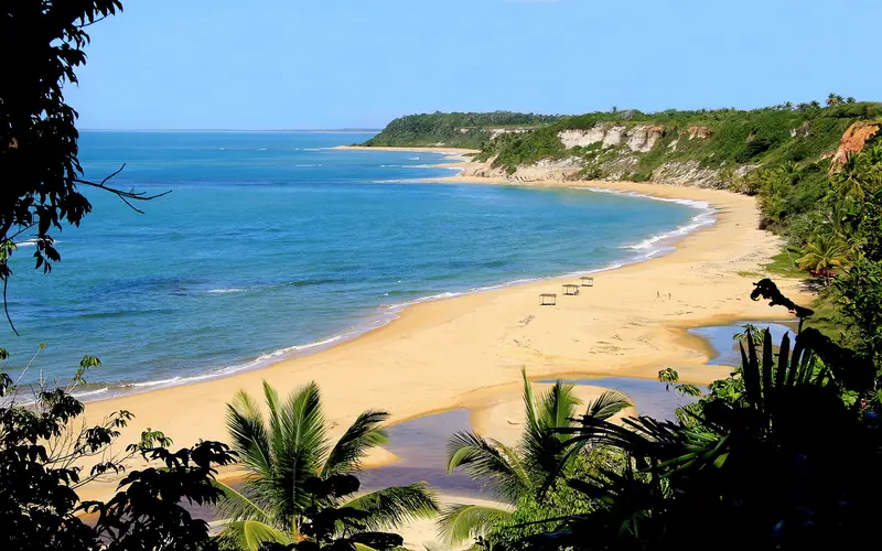 Enseada de areia clara vista do alto, entre mata e falésias brancas e avermelhadas, com o mar azul e um riacho na areia