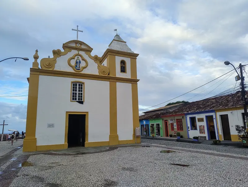 Igreja branca com detalhes amarelos numa praça de pedras portuguesas, com casinhas coloridas ao lado