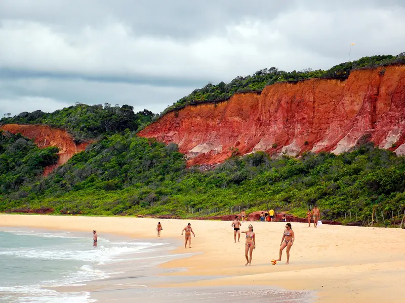 Falésias avermelhadas cobertas de mata no alto, sobre uma praia larga de areia clara