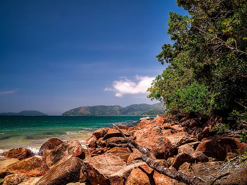 Costão de pedras avermelhadas e mata na Praia do Lázaro, com mar verde e morros ao fundo