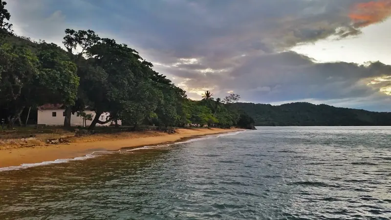 Praia do Presídio no fim da tarde, com areia dourada, árvores grandes e uma casa branca na orla