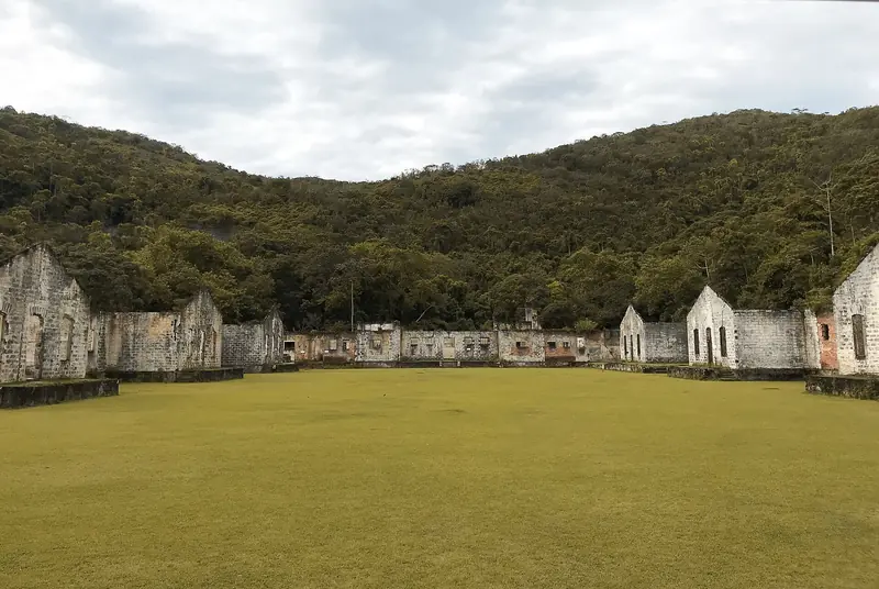 Pátio gramado do antigo presídio da Ilha Anchieta, cercado por pavilhões de pedra em ruínas e morro de mata