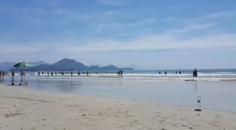 Praia das Toninhas com banhistas na beira da água, guarda-sol e morros ao fundo